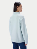 A woman shown from the back wearing a light mint green turtleneck knit sweater and dark blue jeans, against a light gray studio background.