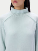 A close-up of a woman wearing a light mint green knit sweater, showing the ribbed turtleneck collar and raglan sleeve seam.