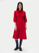A woman modeling a bright red midi dress with three-quarter sleeves and a short button placket, paired with black knee-high boots, against a light gray studio background.