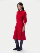 A model wearing a bright red midi dress with three-quarter sleeves and a mock neck, paired with black knee-high boots, against a light gray studio background.