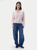 A model stands against a plain light gray studio background wearing a pale pink long-sleeve knit top, loose blue jeans, and dark shoes. _delicate-pink