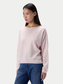 A woman modeling a light pink knit sweater with a wide boat neckline, paired with blue jeans, against a plain light gray studio background. _delicate-pink