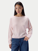 A woman modeling a pale pink long-sleeve knit top with a wide boat neckline, paired with blue jeans, against a light gray studio background.