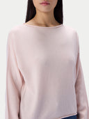 A close-up of a model wearing a pale pink knit sweater with a wide boat neckline and long sleeves, set against a plain light gray background.