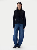 A model stands against a light gray studio background wearing a black long-sleeve mock-neck knit top with a central button placket, paired with loose blue jeans and black shoes.
