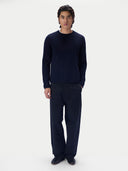 A man wearing a navy blue long-sleeve crewneck sweater, dark trousers and dark shoes, standing against a light gray studio background.