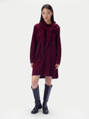 A model wearing a long-sleeved burgundy wrap dress with a large bow detail at the collar, paired with black knee-high leather boots, against a plain light gray background.