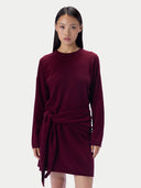 A model wearing a long-sleeve crewneck knit dress in deep burgundy, styled with a self-tie belt at the waist, set against a plain light gray background.