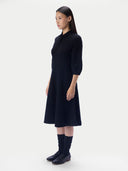 A woman modeling a dark navy midi dress with three-quarter sleeves and a high mock neck, paired with dark socks and shoes, against a plain light gray studio background.