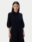 A model wearing a black long-sleeve knit polo dress with three-quarter puff sleeves and a three-button collar, photographed against a solid light gray background.