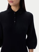 A close-up of a model wearing a black knit polo shirt with three-button placket and three-quarter sleeves against a light gray background.