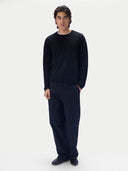 A man wearing a black long-sleeve crewneck sweater, dark trousers and dark shoes, standing against a light gray studio background.