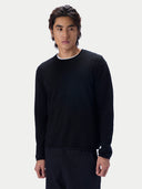 A man wearing a black long-sleeve crewneck knit sweater over a white t-shirt and dark trousers, standing against a light gray studio background.