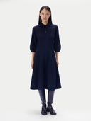 A model stands against a light gray studio background wearing a dark navy blue midi dress with three-quarter sleeves, a polo-style collar, and a buttoned neckline, paired with dark leather boots.