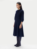 A model is shown in profile wearing a dark navy blue knit dress with three-quarter sleeves and a high mock neck, paired with dark leather knee-high boots, against a plain light gray studio background.