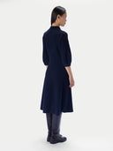 A model shown from the back wearing a navy blue knit dress with elbow-length sleeves, paired with dark leather boots, against a plain light gray studio background.