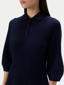 A close-up of a woman wearing a dark navy blue knit polo sweater with three-quarter sleeves and a buttoned collar, shown against a light gray studio background.
