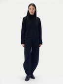A model stands against a plain light gray studio background wearing a matching black knit outfit consisting of a mock-neck long-sleeve top with a front zipper and braided detail, and wide-leg pants.