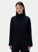 A model wearing a black long-sleeve knit top with a high mock neck and a central braided detail, paired with black pants, against a light gray studio background.
