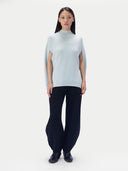 A model stands against a plain light gray studio background wearing a pale icy blue mock-neck knit top with dramatic side slits and long draped sleeves, paired with dark wide-leg pants and black shoes.