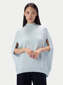 A model wearing a light blue knit poncho sweater with side cutouts, paired with black pants, against a solid light gray background.
