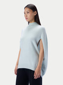 A woman modeling a light blue knit mock-neck cape-sleeve top with side cutouts, paired with black pants, against a plain light gray studio background.