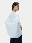 A model shown from a side-back angle wearing a light blue knit poncho-style sweater over a dark navy bottom, set against a plain light gray studio background.