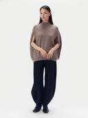 A model stands against a light gray studio background wearing a taupe-brown knit cape-sleeve mock-neck sweater and dark navy wide-leg trousers.