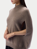 A woman wearing a taupe brown knit sweater with a mock neck and wide batwing sleeves, shown from a side profile against a light gray studio background.