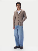 A man wearing a taupe knit blazer-style cardigan over a white t-shirt, wide-leg light blue jeans and dark brown suede shoes, standing against a light gray studio background.