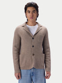 A man wearing a taupe knit blazer-style cardigan over a white t-shirt with light blue jeans, standing against a light gray studio background.