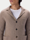 A close-up of a man wearing a taupe knit blazer-style cardigan over a white t-shirt, showing the notched lapel, dark buttons and chest pocket.