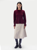 A model stands against a light gray studio background wearing a long-sleeve deep burgundy knit top, a light beige A-line skirt, dark navy boots, and black socks.