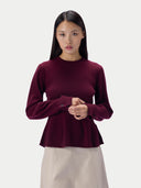 A woman modeling a long-sleeve crewneck knit sweater in deep burgundy, paired with a light beige skirt, against a plain light gray studio background.