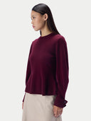 A woman shown in side profile wearing a long-sleeve crew-neck knit sweater in deep burgundy, paired with light beige trousers, against a plain light gray studio background.