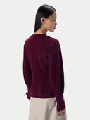 A woman shown from the back wearing a long-sleeve knit sweater in deep burgundy, paired with light beige trousers, against a plain light gray studio background.