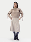 A model stands against a light gray studio background wearing a beige ribbed knit sweater with dramatic shoulder tie details, a light beige midi skirt, dark navy boots, and black leggings.