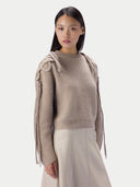 A model wearing a beige ribbed knit sweater with unique knotted shoulder details and long hanging fringe, paired with a light beige skirt, against a solid light gray background.