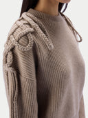 Close-up of a person wearing a taupe ribbed knit sweater with chunky braided rope details across the shoulders and draped down one sleeve, against a plain light background.