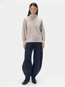 A model stands against a light gray studio background wearing a light beige knit turtleneck sweater and dark navy wide-leg trousers.