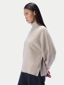 A woman in side profile wearing a light beige knit mock-neck sweater and dark charcoal trousers, photographed against a clean off-white studio background.