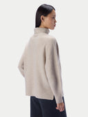 A woman shown from the back wearing a light beige knit turtleneck sweater and dark trousers, photographed against a clean light gray studio background.