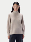 A woman modeling a light beige turtleneck sweater against a plain light gray studio background.