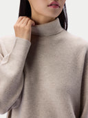 Close-up of a model wearing a light beige knit turtleneck sweater against a plain light background.