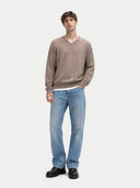 A man modeling a taupe brown V-neck knit sweater, light blue jeans, and black leather shoes against a light gray studio background.