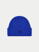 A bright royal blue ribbed knit beanie with a folded cuff, featuring a small black embroidered geometric logo in the center of the cuff, set against a light gray background.