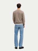 A man viewed from behind wearing a taupe crewneck sweater, light blue jeans, and black shoes, standing against a light gray studio background.