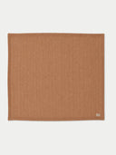 A square woven placemat in a warm camel brown color, featuring a subtle vertical ribbed texture and a small white brand tag in the bottom right corner, displayed on a clean off-white background.
