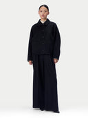 A model stands against a light gray studio background wearing a dark navy or near-black long-sleeve button-front jacket with large front patch pockets, paired with matching wide-leg pants.