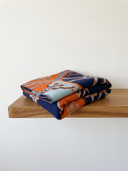 A folded patterned scarf or throw blanket with navy blue, orange, and light blue sections, displayed on a wooden shelf against a plain white wall.